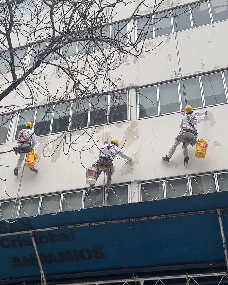 Tres operarios de Pintura Express trabajando en silleta sobre la fachada de un edificio, con arnés, casco y línea de vida